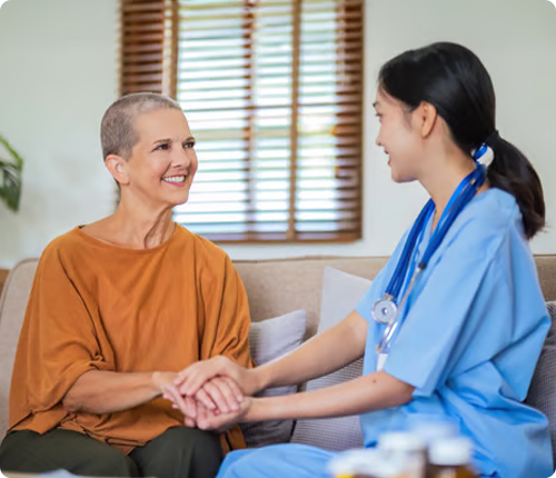 An Asian nurse in a blue uniform gently holds the hand of a smiling senior Caucasian woman with a shaved head, who is wearing an orange shirt. They are sitting on a couch in a home setting.