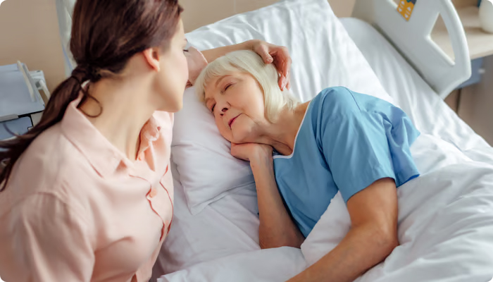 A young woman in a peach shirt sits by a hospital bed, holding the hand of an elderly woman with white hair who is lying down and resting.