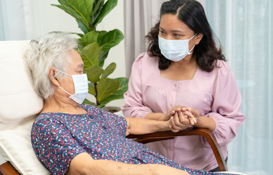 A family caregiver wearing a protective face mask holds the hand of an elderly woman resting in a chair, illustrating the emotional support and personal connection central to hospice care.