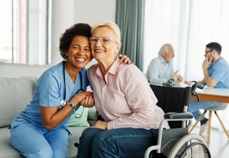 A smiling female hospice nurse in blue scrubs sits on a sofa, warmly embracing a senior woman in a wheelchair who is smiling back. In the background, a male healthcare provider assists an elderly man.