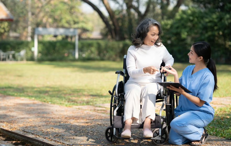 A healthcare professional in blue scrubs kneeling beside an elderly woman in a wheelchair in an outdoor park setting, discussing information on a clipboard.