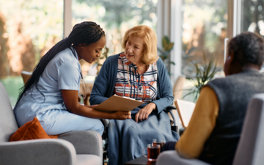 An African American nurse in light blue scrubs reviews a clipboard with a senior woman in a wheelchair in a bright nursing home common area.