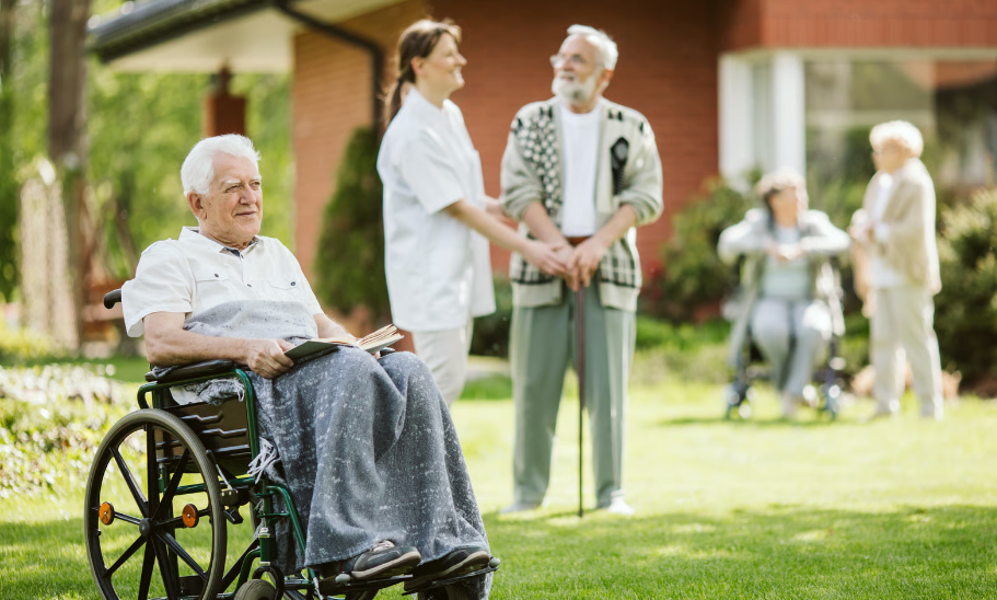An elderly man with white hair sits in a wheelchair with a blanket over his legs holding a book on a green lawn, while a female nurse and other senior residents talk in the blurred background outside a residential facility.