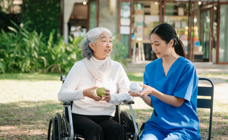 An Asian female nurse in blue scrubs sits outdoors next to an elderly Asian woman in a wheelchair, explaining the label on a white medicine bottle while the senior woman holds a green apple.
