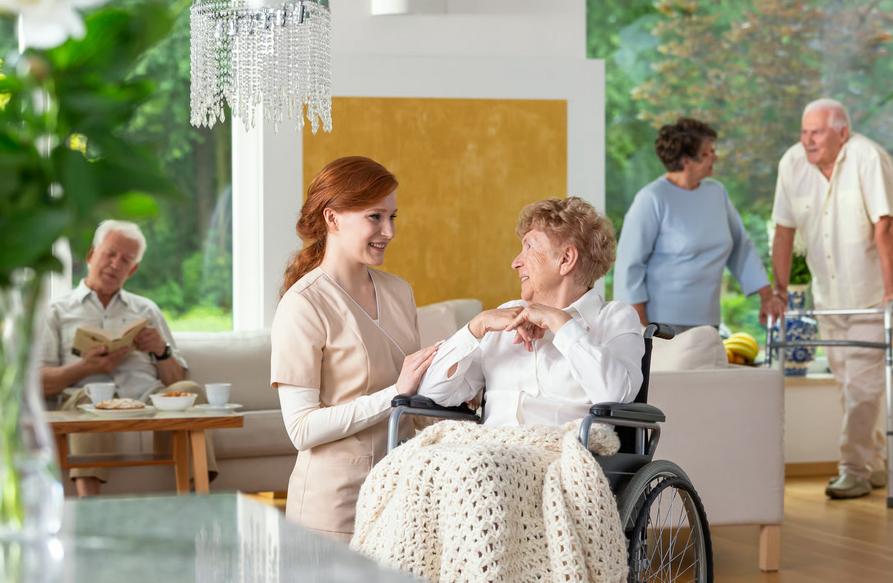 A smiling hospice caregiver talks with an elderly woman in a wheelchair inside a bright, comfortable living space, while other seniors relax in the background. This demonstrates how early hospice care improves comfort and provides a peaceful, supportive environment for patients.