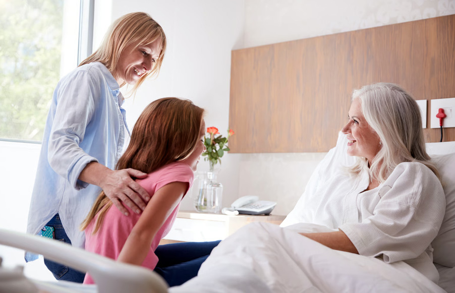 An elderly woman with long white hair smiles warmly from a hospital bed during a comforting visit from her daughter and young granddaughter, showcasing the family connection and emotional support provided through comfort-focused hospice care.