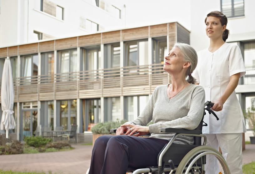 A female nurse in white medical scrubs standing behind and pushing an elderly woman in a wheelchair on an outdoor pathway, both looking up and smiling calmly, representing the early initiation of supportive palliative care for seniors living with a serious illness.