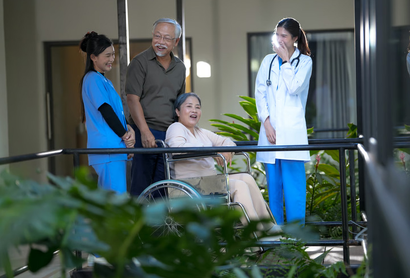 An elderly woman in a wheelchair laughing with her husband, nurse, and doctor on a sunny outdoor patio.