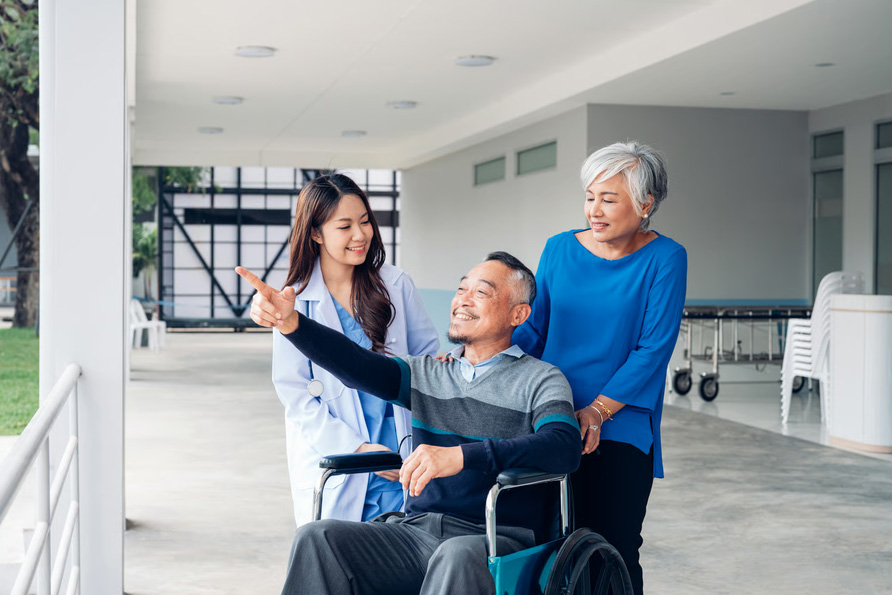An elderly man in a wheelchair pointing outdoors alongside his wife and a smiling nurse at a care facility.