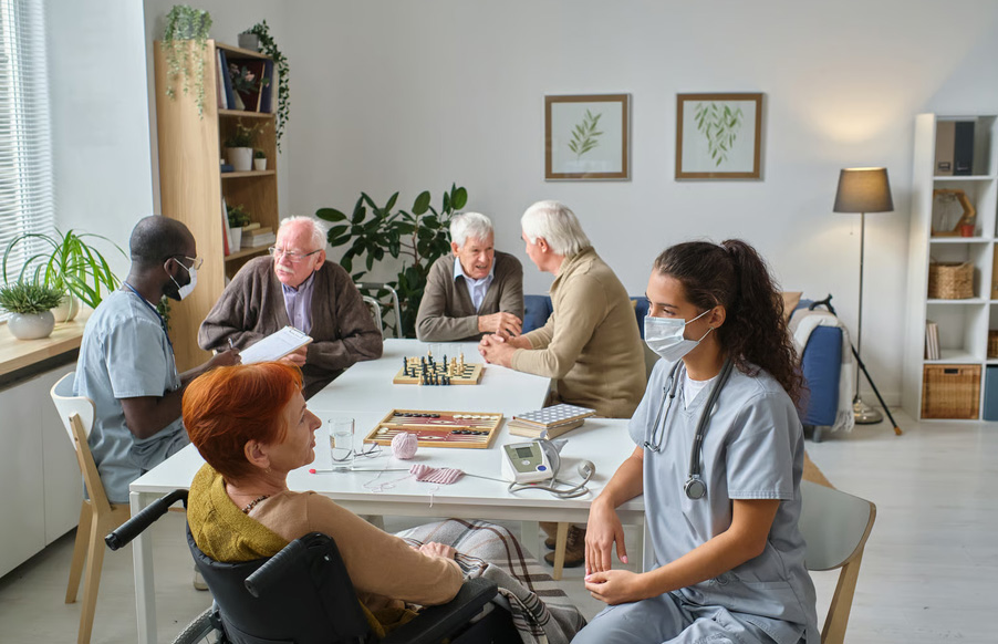 A masked healthcare professional in blue scrubs speaks with a senior woman sitting in a wheelchair, while another masked nurse assists seniors playing a game at a table.