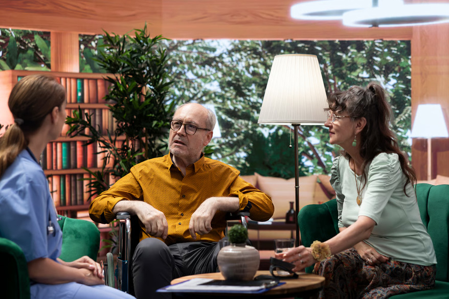 A healthcare professional in blue scrubs sitting in a green chair, discussing a care plan with a senior man in a wheelchair and a woman sitting beside him.