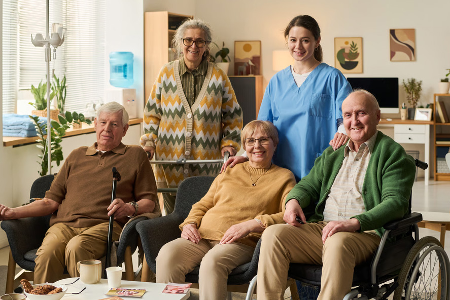 A nurse in blue scrubs stands with four elderly adults sitting in a brightly lit care home living room.
