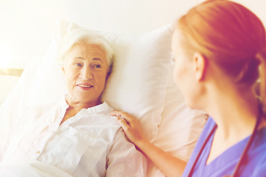 A hospice nurse gently resting a hand on an elderly woman's shoulder as she rests in bed.