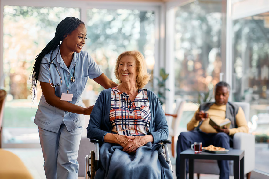 A nurse in scrubs gently resting her hands on an elderly woman's wheelchair while both smile in a sunlit room.