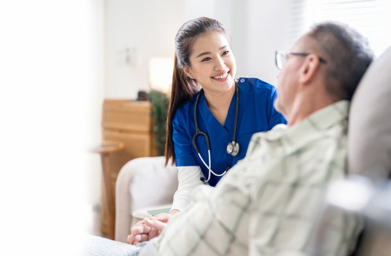 A compassionate hospice nurse in blue scrubs smiling while holding hands with an elderly patient in a home setting.