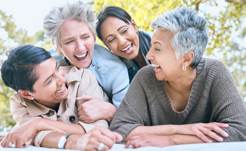 A group of four diverse women lying on a blanket outdoors, laughing together during a respite care break from caregiving.