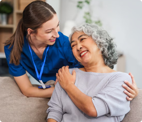 A young female nurse in blue scrubs smiling and leaning in toward a seated senior woman with gray hair who is laughing and looking back at her.