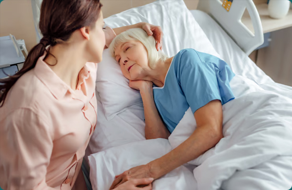 A young woman in a peach shirt sits by a hospital bed, holding the hand of an elderly woman with white hair who is lying down and resting